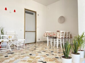 Zona pranzo con pavimento piastrellato, sedie bianche, tavolo con tovaglia floreale e piante in vaso.