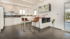 A modern kitchen with white cabinets, a central island, two bar stools, and a dining table. - Villa Red Hacienda with Pool (Vodice)
