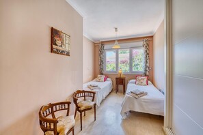 Una camera da letto con due letti singoli, una sedia in legno, un comodino con una lampada e una finestra con tende.