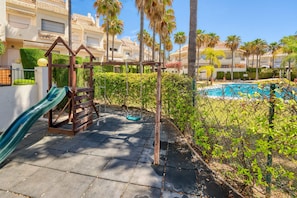 A poolside area with a swing set, a slide, and a clear view of a swimming pool surrounded by palm trees and a hedge.