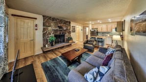 A spacious living room with a stone fireplace, a sofa set, and a kitchen area in the background. - Hi Country Haus Unit 1503 Ground floor 1 bedroom next to the Fraser River (Winter Park)