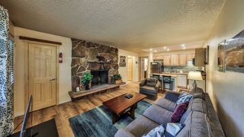 A spacious living room with a stone fireplace, a sofa set, and a kitchen area in the background.