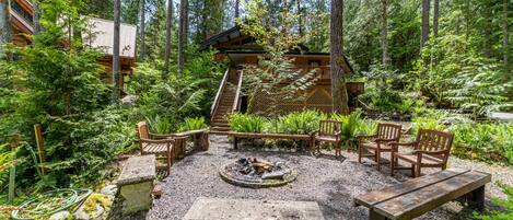A rustic outdoor seating area with wooden benches and a fire pit surrounded by dense forest.