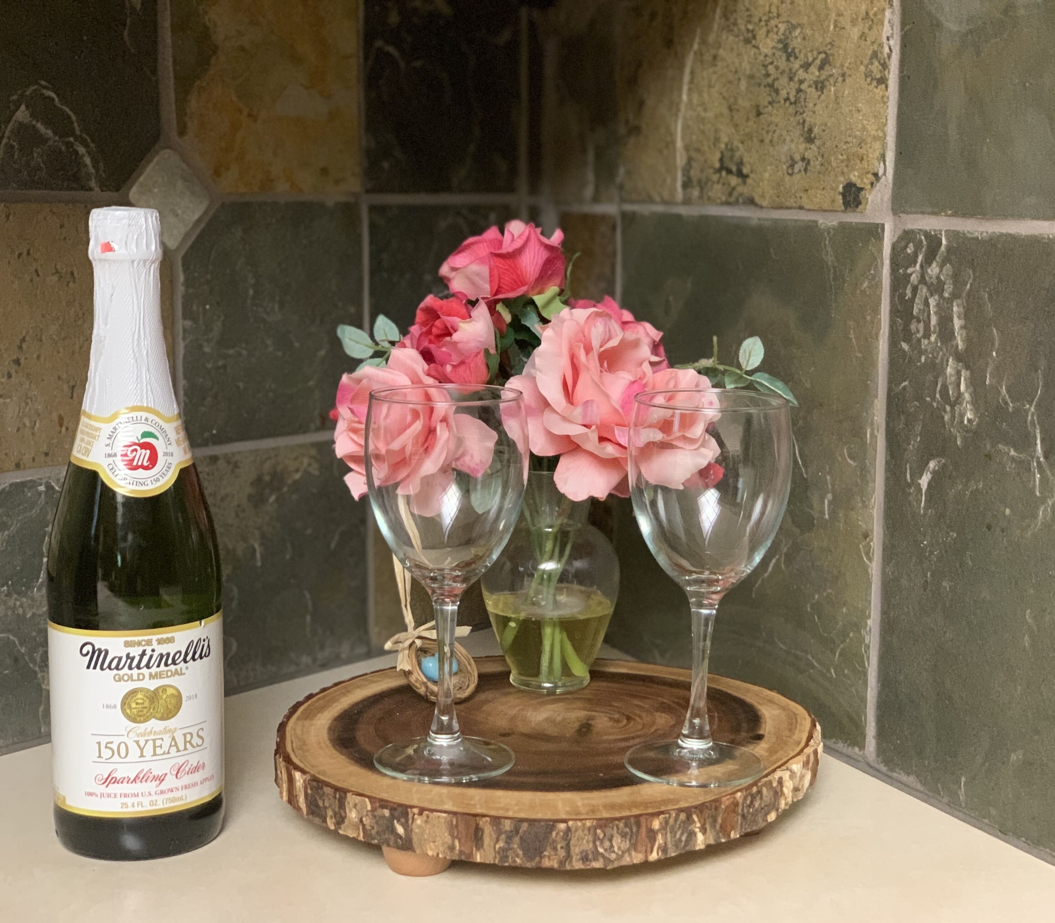 Een fles sprankelende cider, twee wijnglazen en roze bloemen op een houten onderzetter.