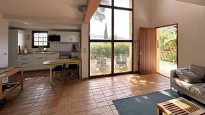 Un vaste espace de vie avec une grande porte-fenêtre coulissante donnant sur un patio, une cuisine avec des meubles blancs et un coin repas comprenant une table et des chaises.