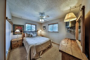 Een slaapkamer met een bed, twee nachtkastjes, een plafondventilator, een raam met zonwering, een lamp, een spiegel en een houten dressoir.