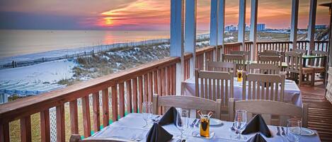 A restaurant with tables set for dining, overlooking a beach with a sunset view.