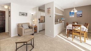 A living room with a sofa, coffee table, and dining area with chairs and a table. There is a kitchen in the background.