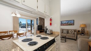 A compact kitchen with a stove, dining table, and chairs. There is a living area with a sofa and a TV. A fire extinguisher is mounted on the wall.