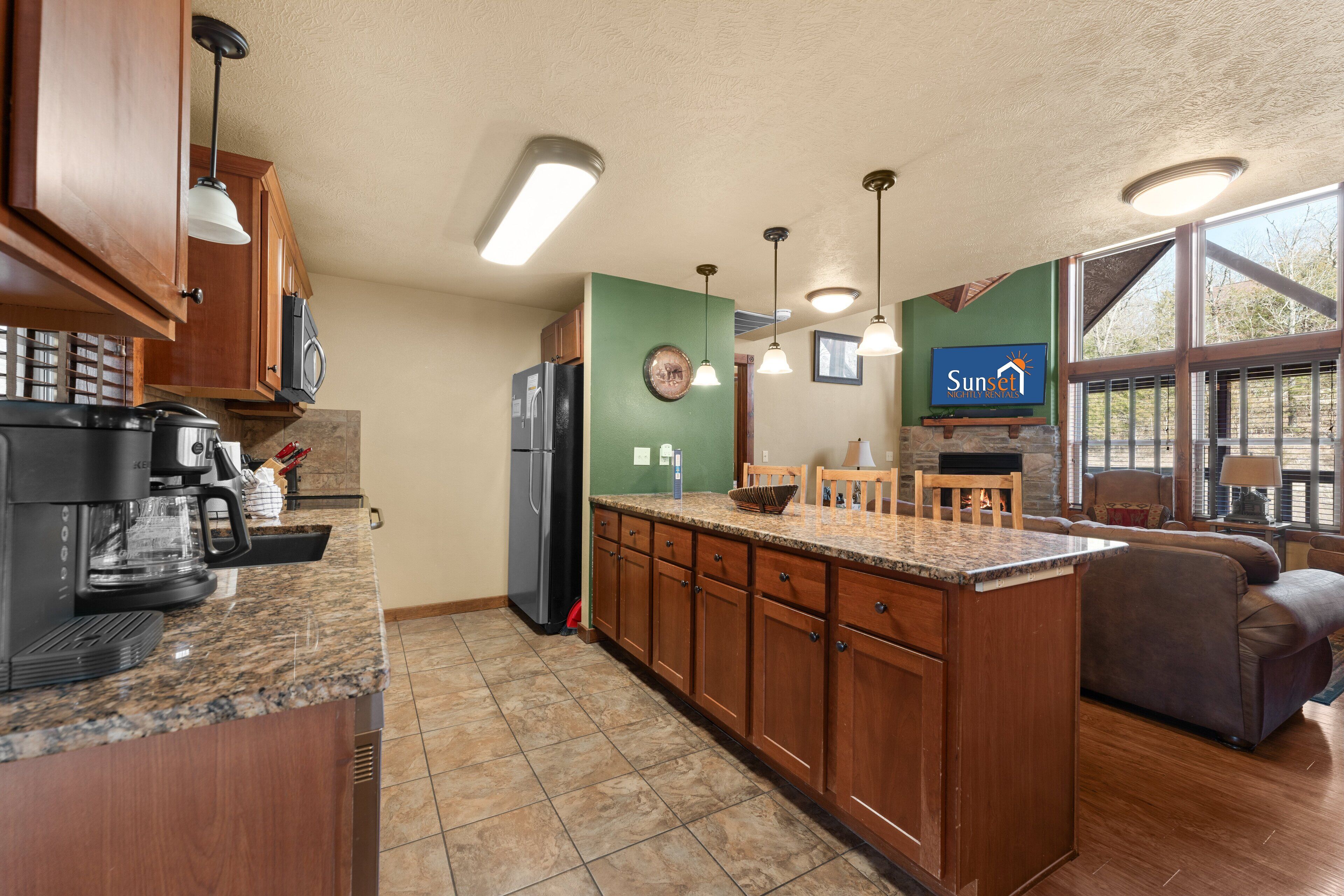 A modern kitchen with granite countertops, wooden cabinets, and a built-in island. There is a refrigerator, microwave, and coffee maker on the counter. The room has a large window providing natural light and a view of the outdoors.