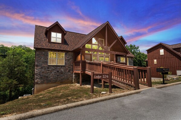 A two-story lodge with a stone facade, a wooden deck, and a clear sky. - Exquisite Decor in this 6 BD Cabin! Super Close to Fun Attractions and Shops! (Branson West)