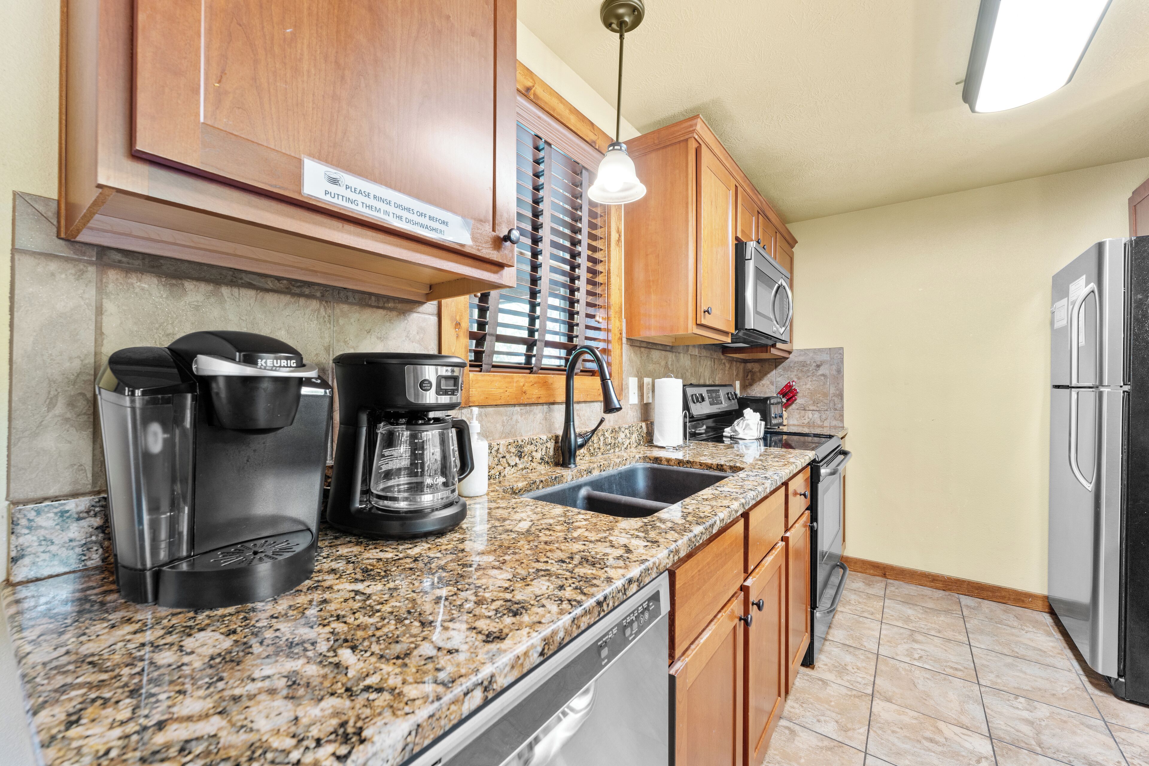 Cocina con encimeras de granito, armarios de madera, refrigerador de acero inoxidable, microondas, cafetera y lavavajillas.