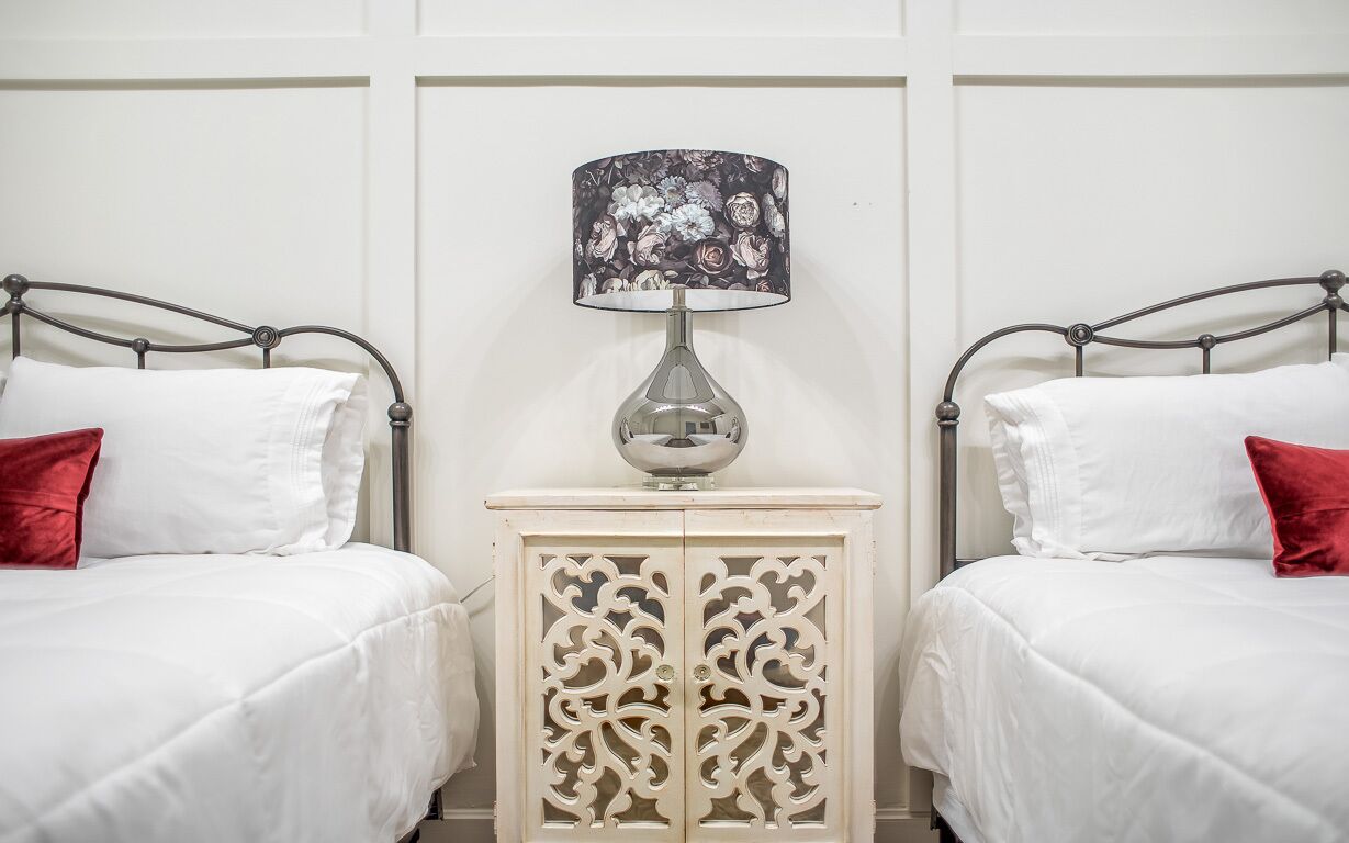 Ein Zimmer mit zwei Betten, jeweils mit weißer Bettwäsche und einem roten Kissen. Eine dekorative Lampe mit floralem Lampenschirm steht auf einer weißen Kommode mit aufwendigen Schnitzereien.