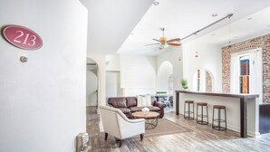 Een moderne woonkamer met een bruine leren bank, een witte fauteuil en een salontafel. Er is een keuken met een bar en krukken. De kamer heeft een hoge plafond met een ventilator en een bakstenen muur.