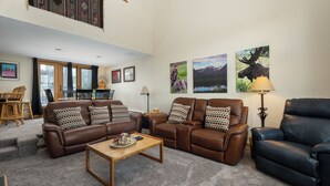 Ein geräumiges Wohnzimmer mit zwei braunen Ledersofas, einem Holztisch und einem schwarzen Ledersessel. An den Wänden befinden sich gerahmte Bilder, eine Lampe und im Hintergrund ein Essbereich.