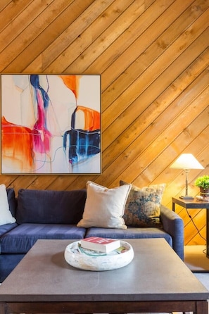 Ein gemütliches Zimmer mit holzverkleideter Schräge, einem blauen Sofa mit Kissen, einem Couchtisch mit einem Buch und einem farbenfrohen, abstrakten Gemälde an der Wand.