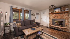 Een gezellige woonkamer met een stenen openhaard, een bank, een salontafel en uitzicht op de natuur door een raam met gordijnen.