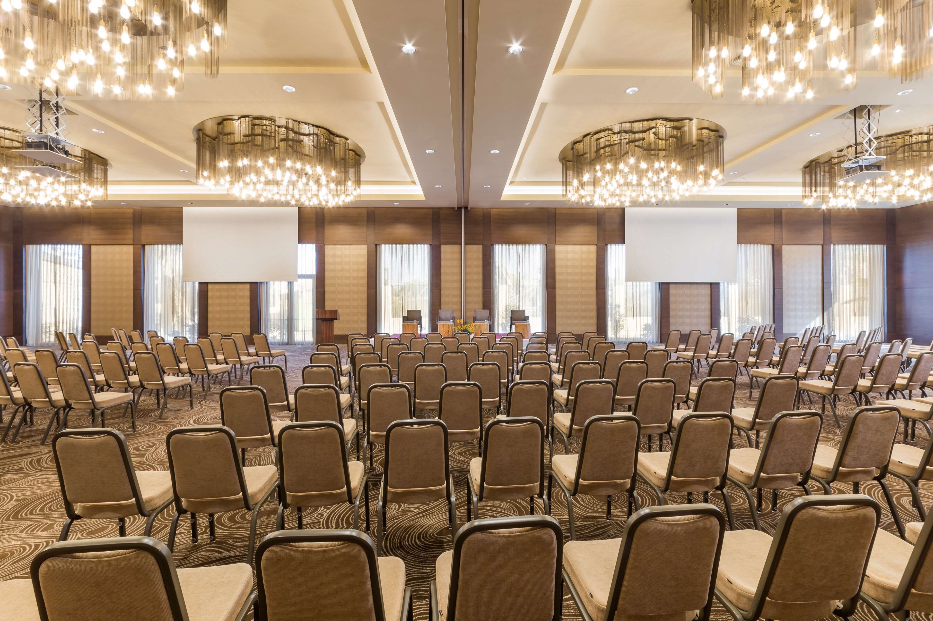 Une salle de conférence avec des rangées de chaises, de grands lustres et une scène équipée d’un écran.