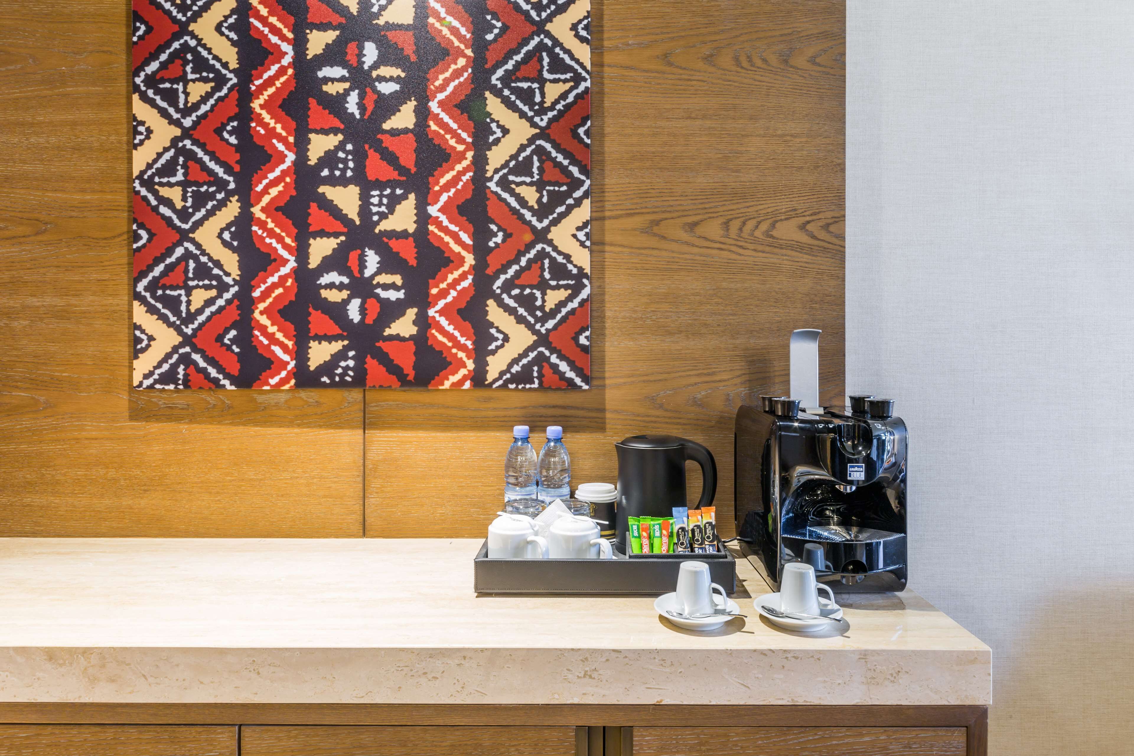 A hotel room with a wooden panel wall, a patterned artwork, a coffee station with a coffee maker, cups, and a tray with water bottles and juice boxes.