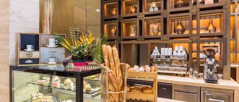 Mostrador de panadería con pasteles, pan, una máquina de café y una planta decorativa.