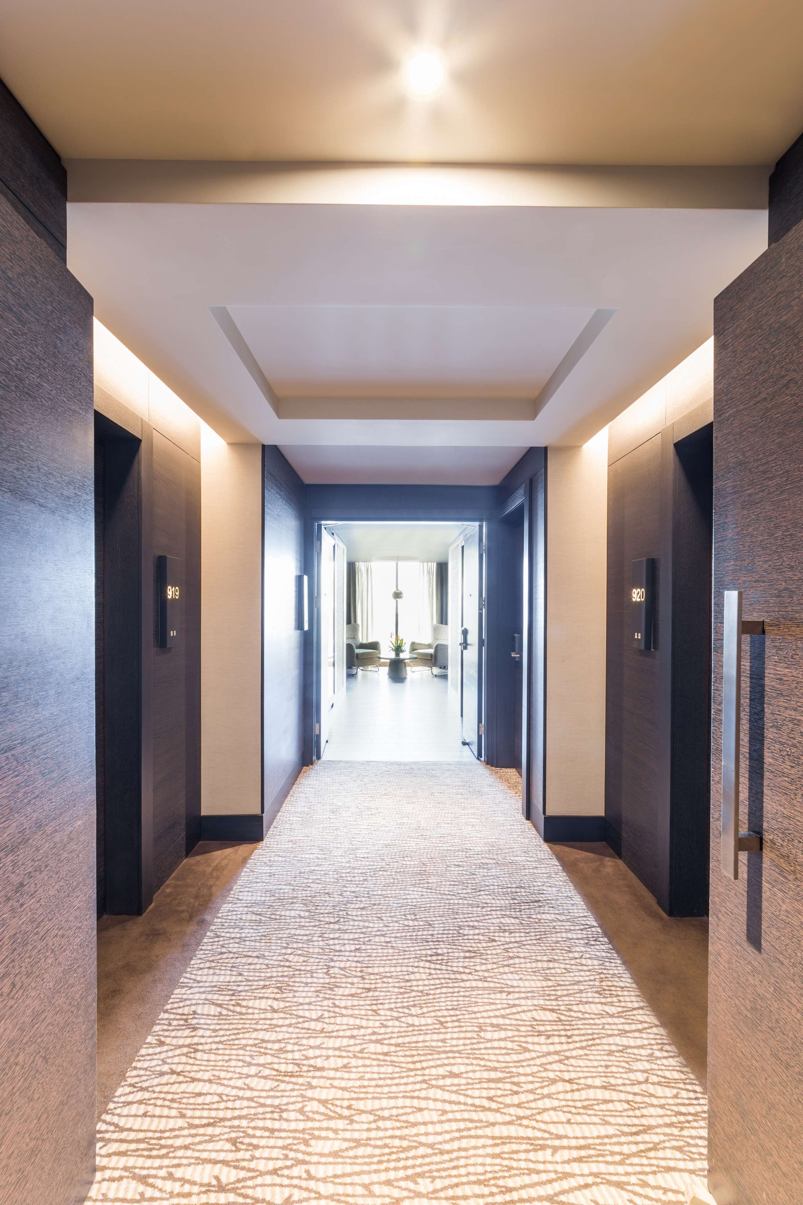 Un pasillo moderno de hotel con alfombra estampada, paredes oscuras y placas con números iluminados en las puertas.