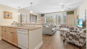 A modern kitchen with a dishwasher, wooden cabinets, and a large island. There is a dining area with a sofa and a television.