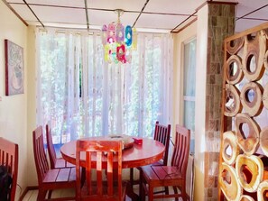 A dining area with a round table and six wooden chairs, large windows with sheer curtains, and a decorative wooden panel on the right wall. - Hwan Dynasty Suite Elnido (El Nido)