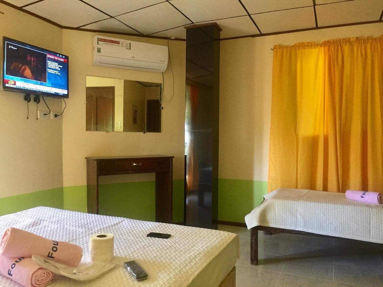 Habitación de hotel con cama, mesita de noche con teléfono y control remoto, espejo, televisión y ventana con cortinas amarillas.