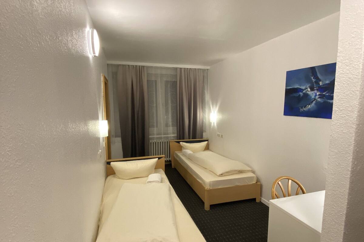 Habitación de hotel con dos camas individuales, un escritorio pequeño, una silla, una ventana con cortinas y un cuadro en la pared.