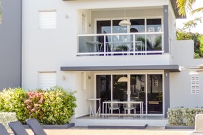 A modern two-story building with a balcony, a dining area with a table and chairs, and a small garden area with plants and a seating bench. - Beluga Apartments (Willemstad)