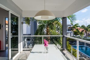 A balcony with a white table, chairs, a vase with pink flowers, and a view of a swimming pool and palm trees. - Beluga Apartments (Willemstad)