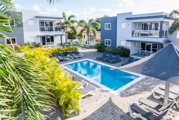 A modern pool area with lounge chairs and a clear blue pool, surrounded by buildings with balconies and a tropical garden. - Beluga Apartments (Willemstad)