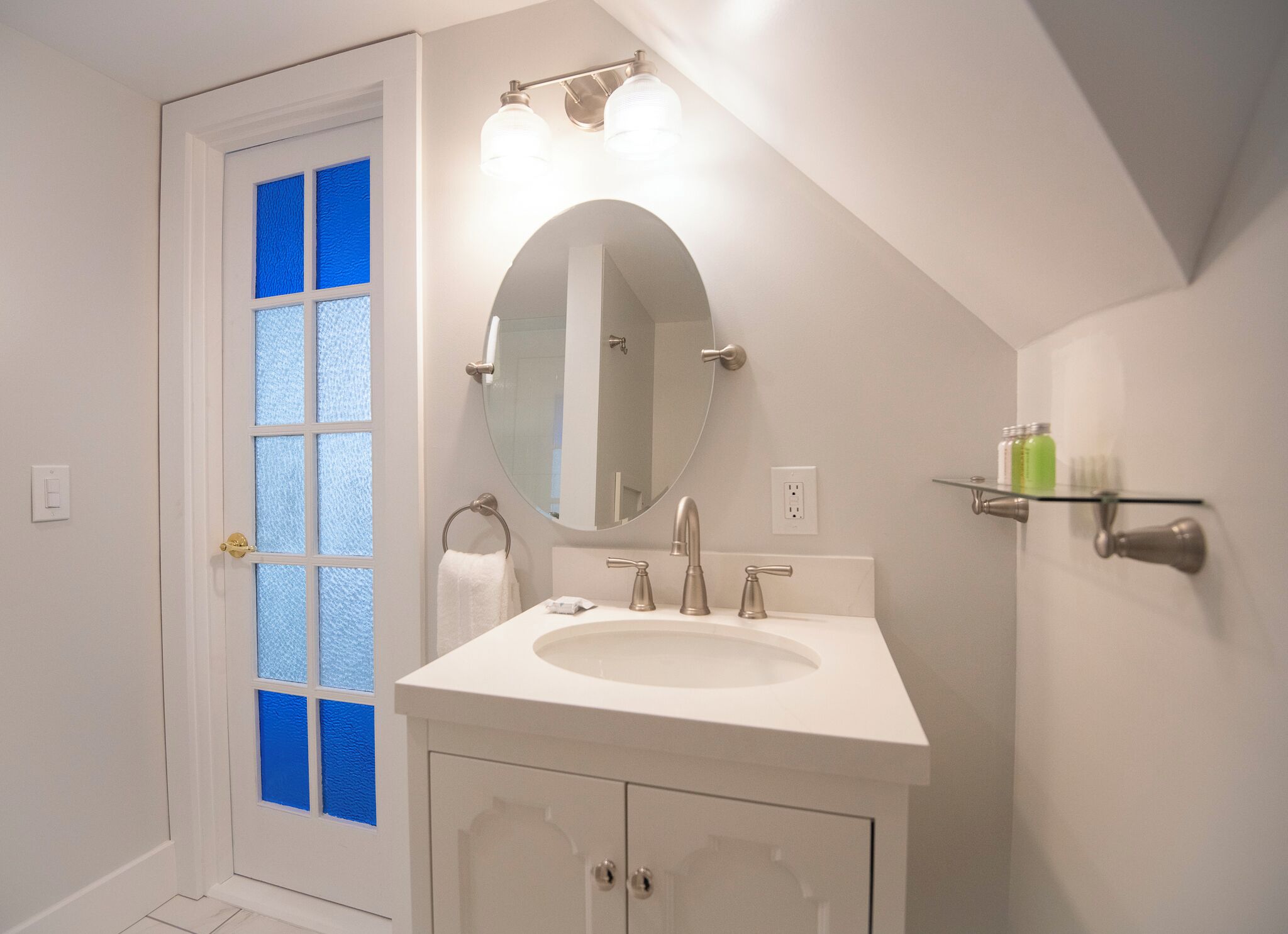 A bathroom with a white sink, a round mirror, and a glass door with blue tint.