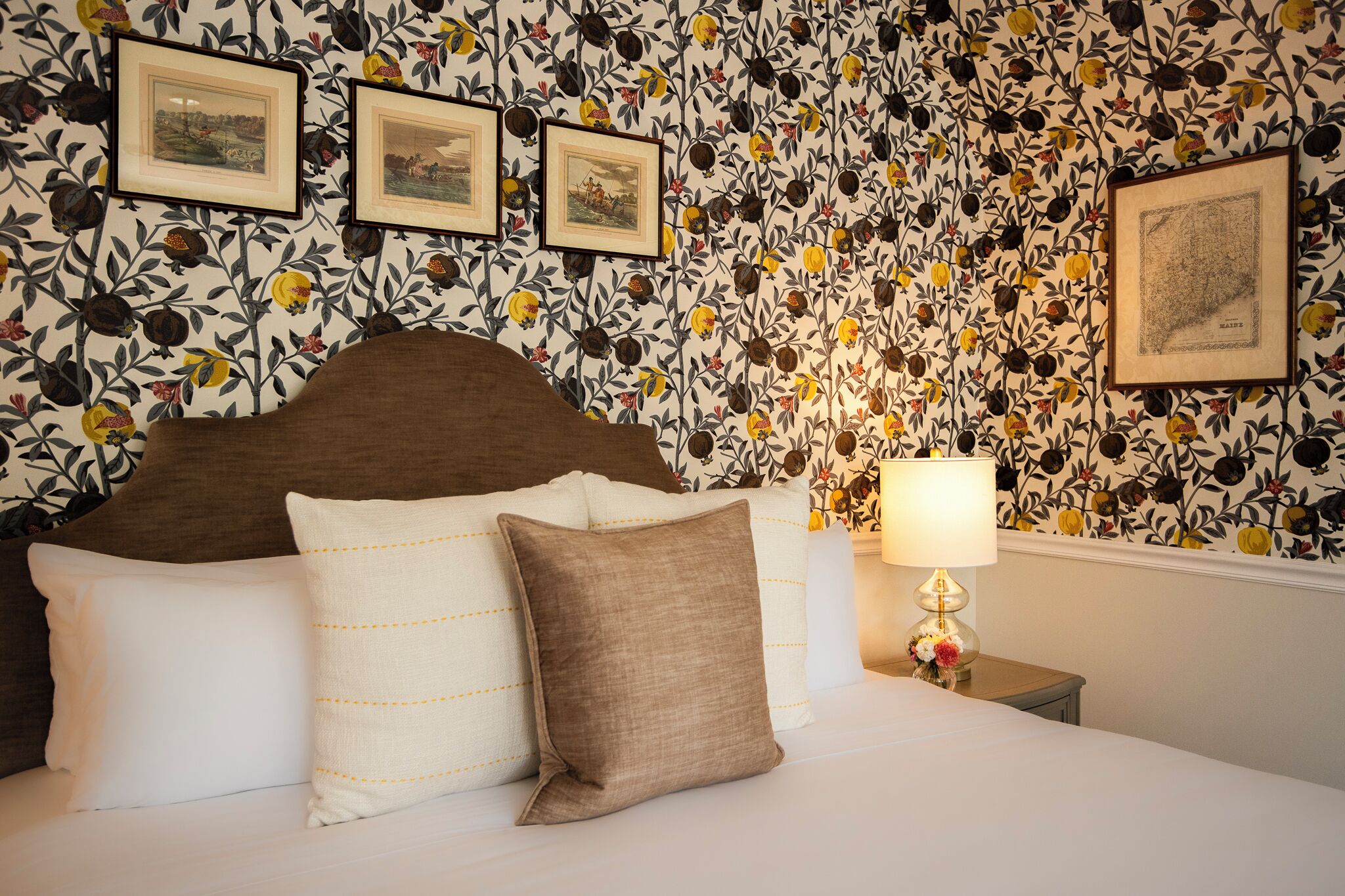 A neatly made bed with white and brown pillows, a wooden headboard, and a floral wallpaper pattern.