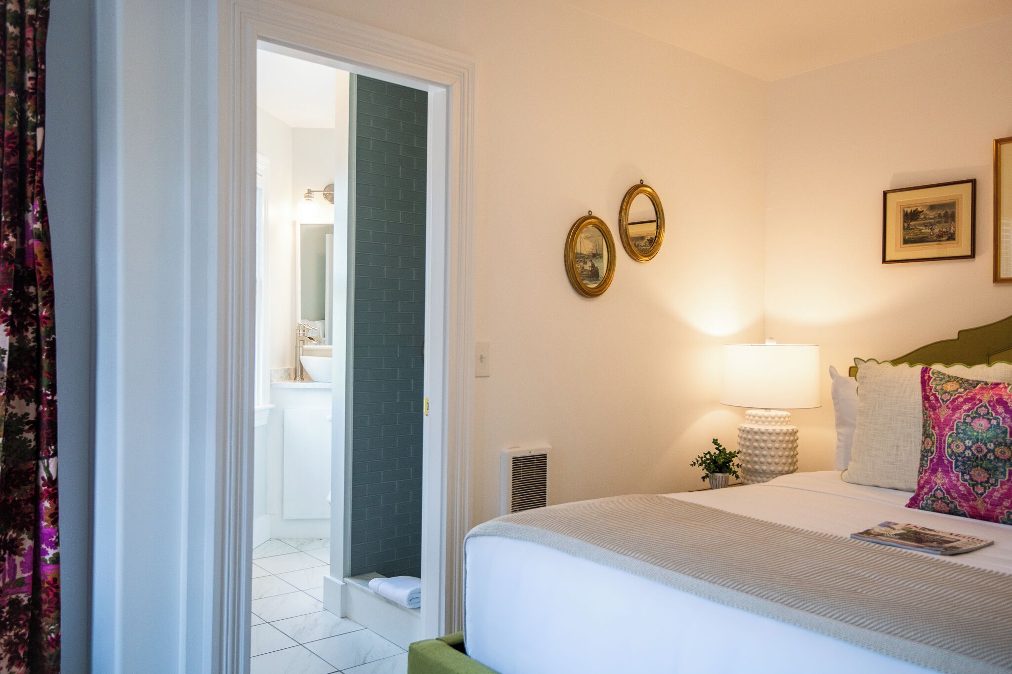 Habitación de hotel con cama, mesita de noche, lámpara, dos cuadros enmarcados y un baño visible a través de una puerta abierta.