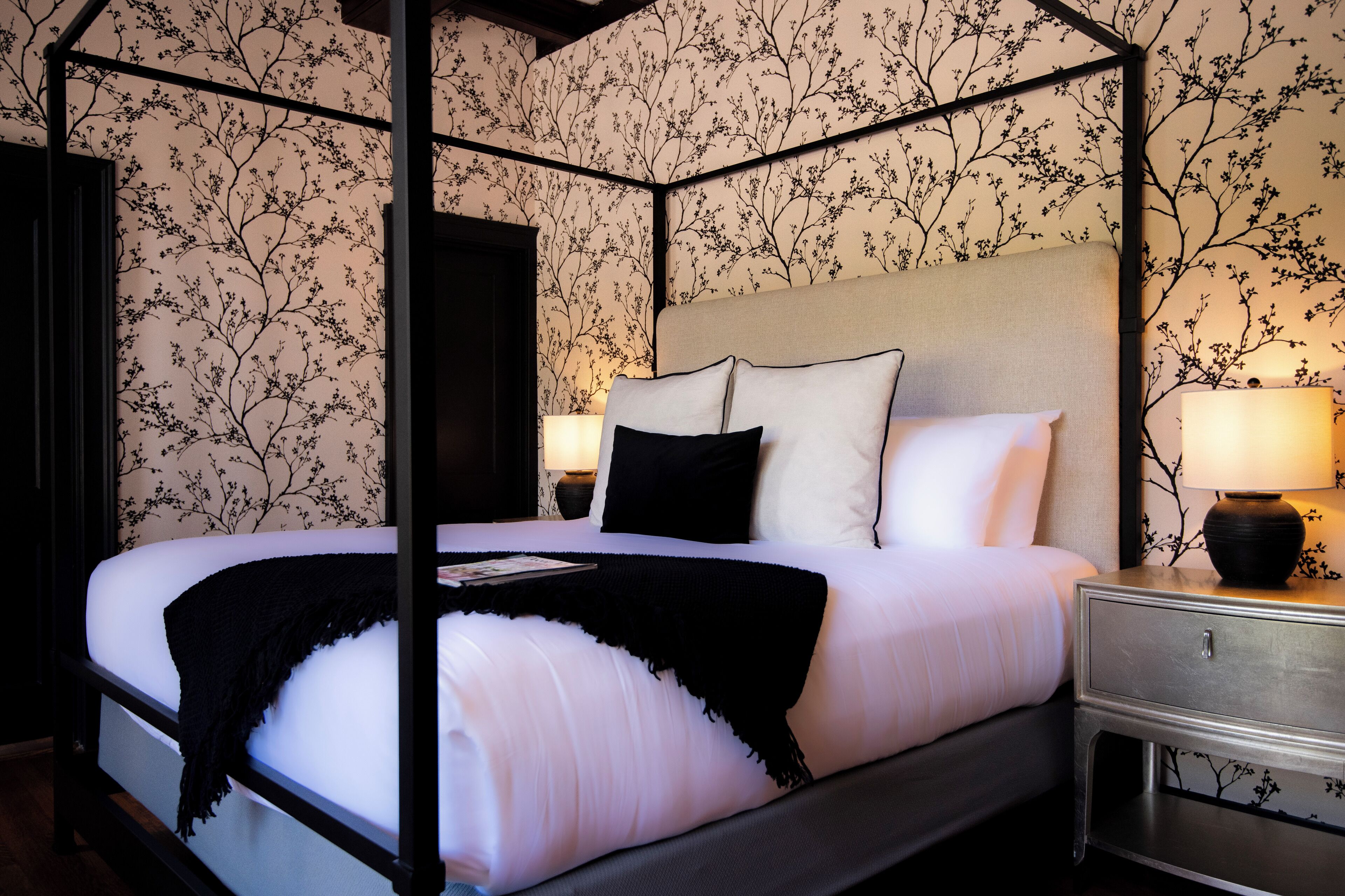 Una cama con dosel y cuatro columnas con ropa de cama blanca y almohadones negros, una mesita de noche con una lámpara y papel tapiz floral.