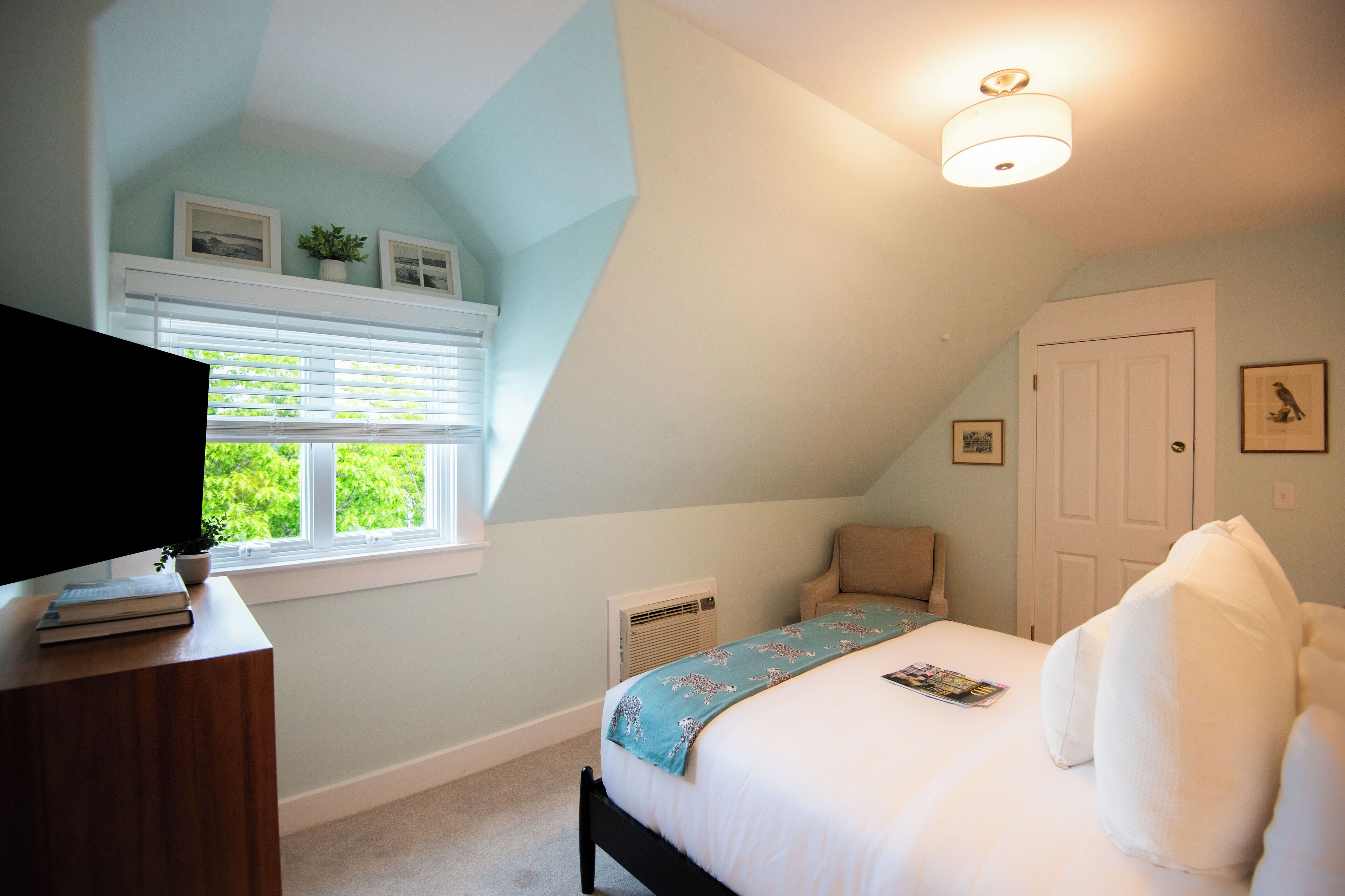 A bedroom with a bed, a chair, a desk with a TV, a window with blinds, and a ceiling light.