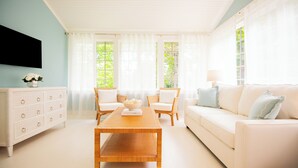 Un salón luminoso con un sofá blanco, dos sillones blancos, una mesa de centro de madera y un mueble blanco con cajones.