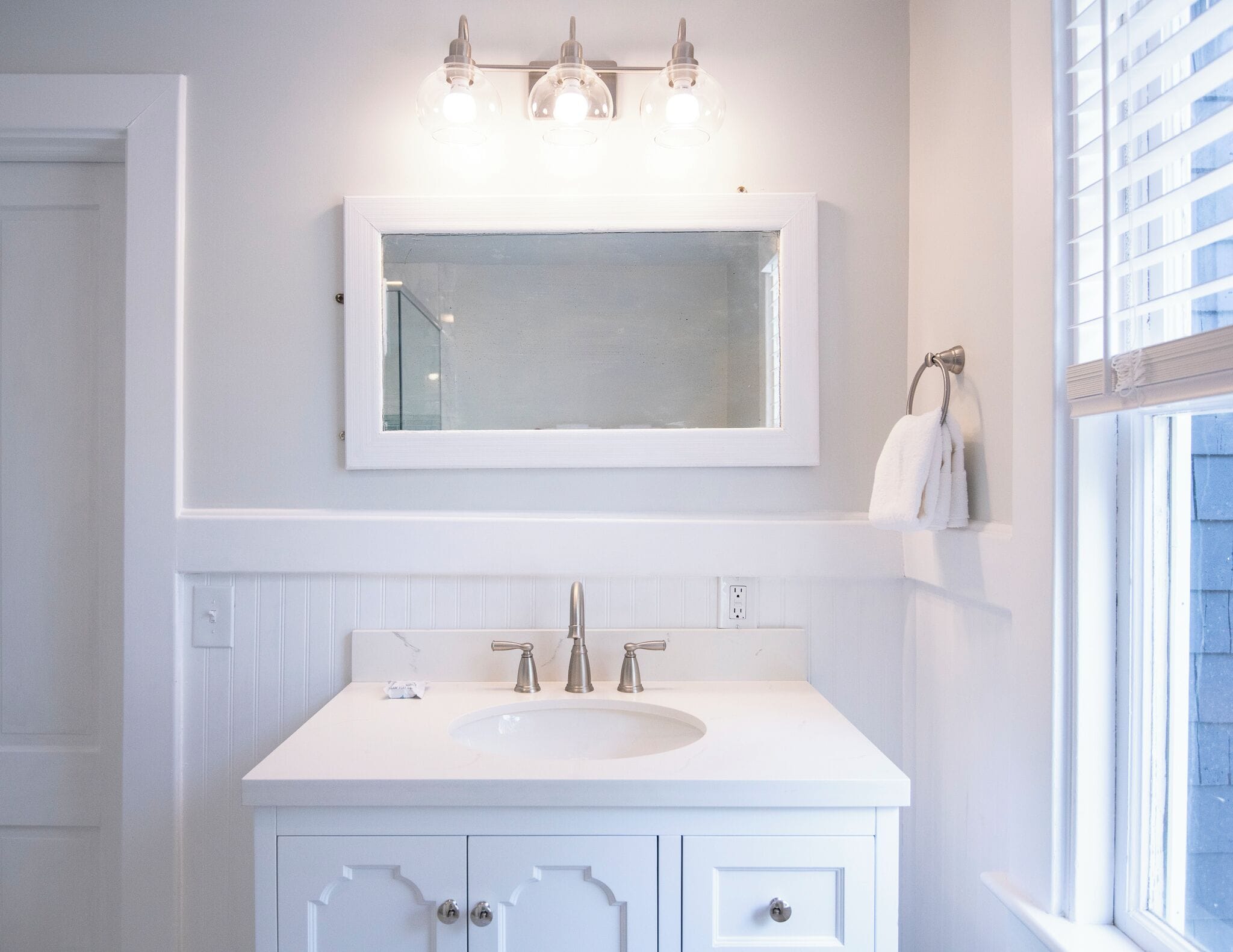Une salle de bain avec un meuble-lavabo blanc, un grand miroir et une fenêtre avec des stores.