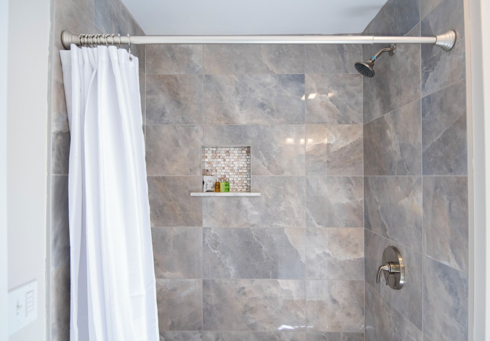 Eine Dusche mit weißem Duschvorhang, einer Handbrause und einem Ablagefach mit Toilettenartikeln.