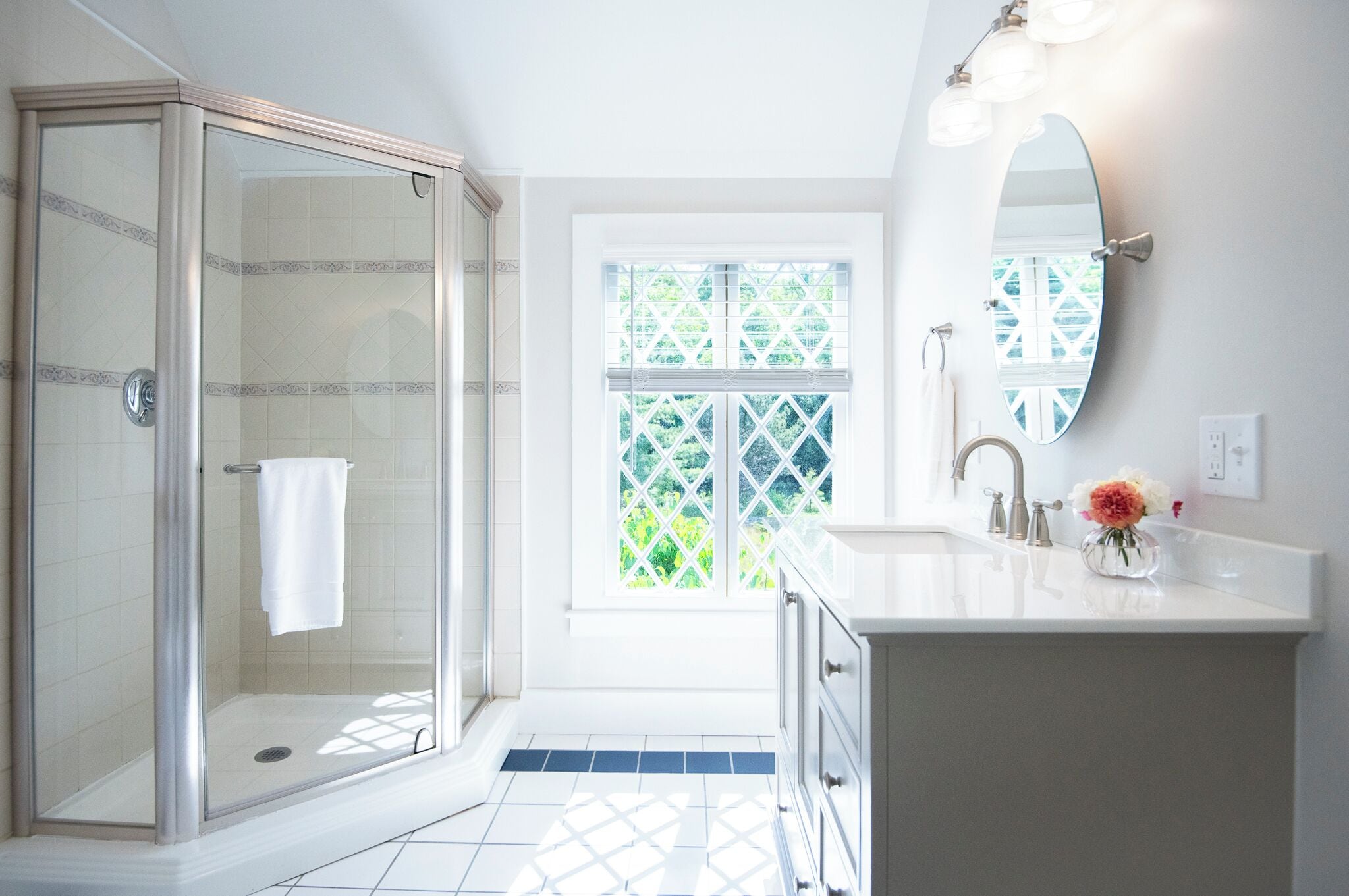 A modern bathroom with a glass shower enclosure, a large window with a view of greenery, and a white vanity with a mirror and a vase of flowers.