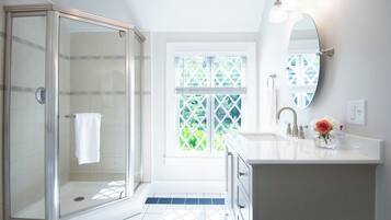 A modern bathroom with a glass shower enclosure, a large window with a view of greenery, and a white vanity with a mirror and a vase of flowers.