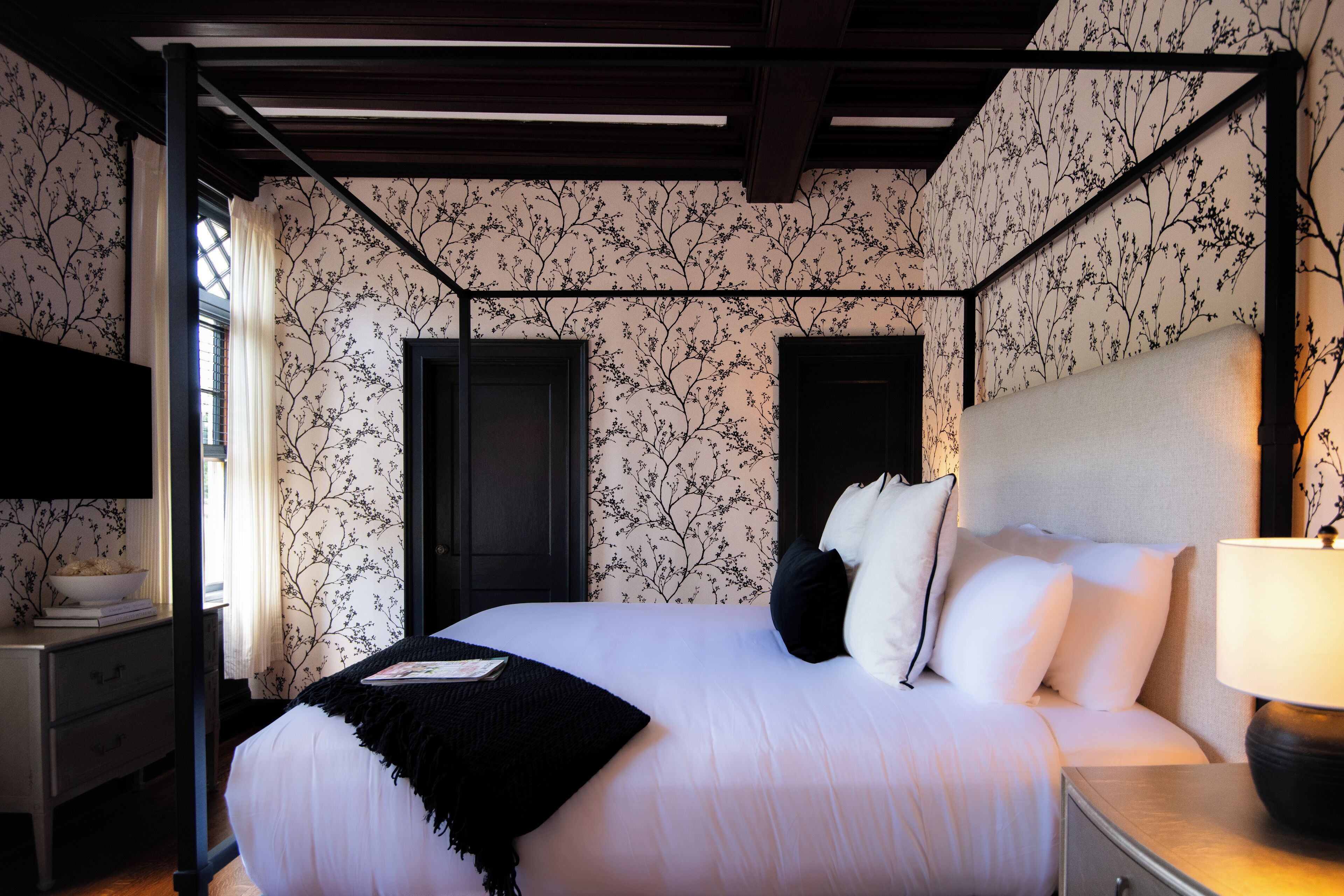A bedroom with a four-poster bed, a bedside table with a lamp, and a window with curtains.
