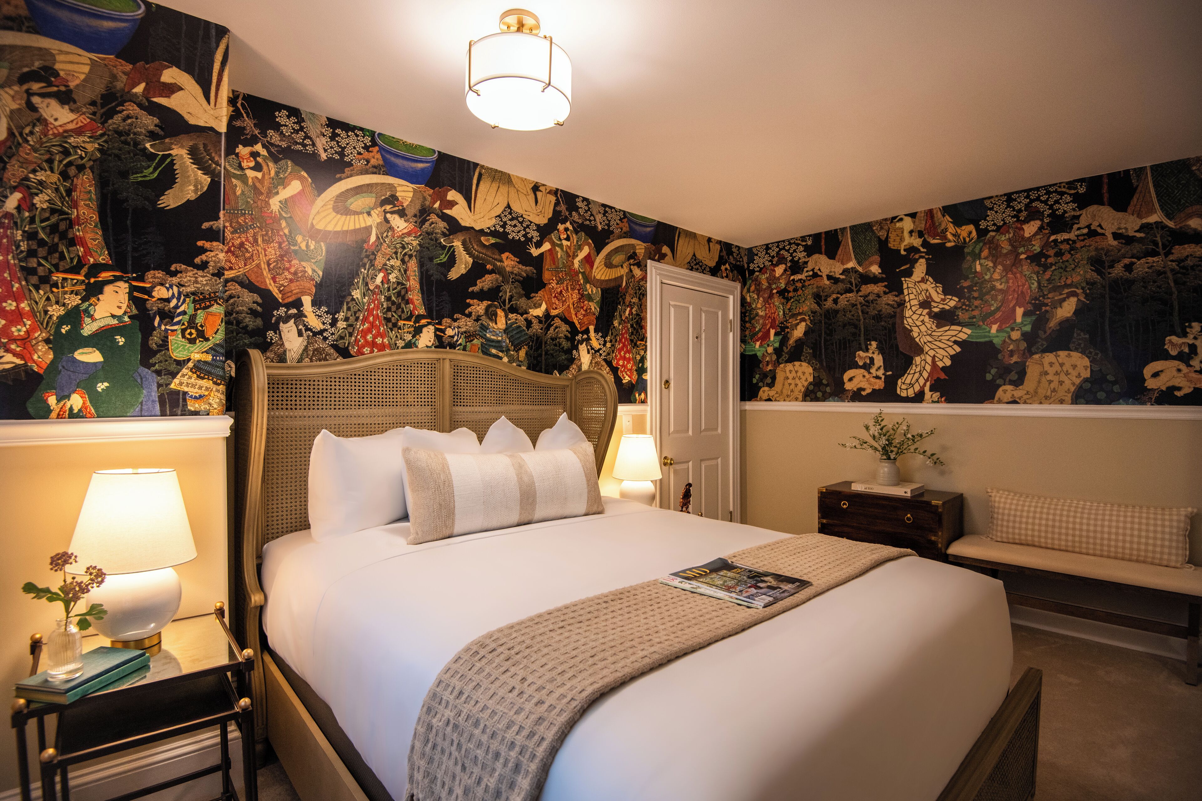 A hotel room with a large bed, a bedside table with a lamp, a bench, and a wall with traditional artwork.
