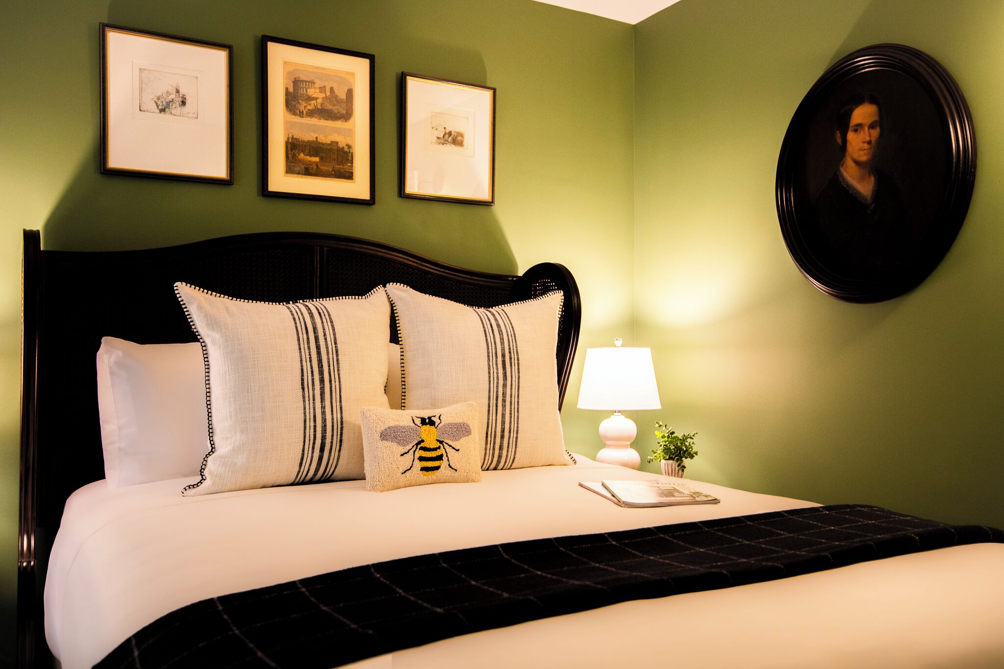 A neatly made bed with a black headboard, white pillows, and a bee-themed pillow. There are three framed pictures on the wall above the bed.
