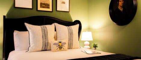 A neatly made bed with a black headboard, white pillows, and a bee-themed pillow. There are three framed pictures on the wall above the bed.