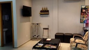Una sala moderna con televisor de pantalla plana, una mesita de centro con revistas, una pequeña mesa con una silla, y un área de estar con sillones de estampado floral.