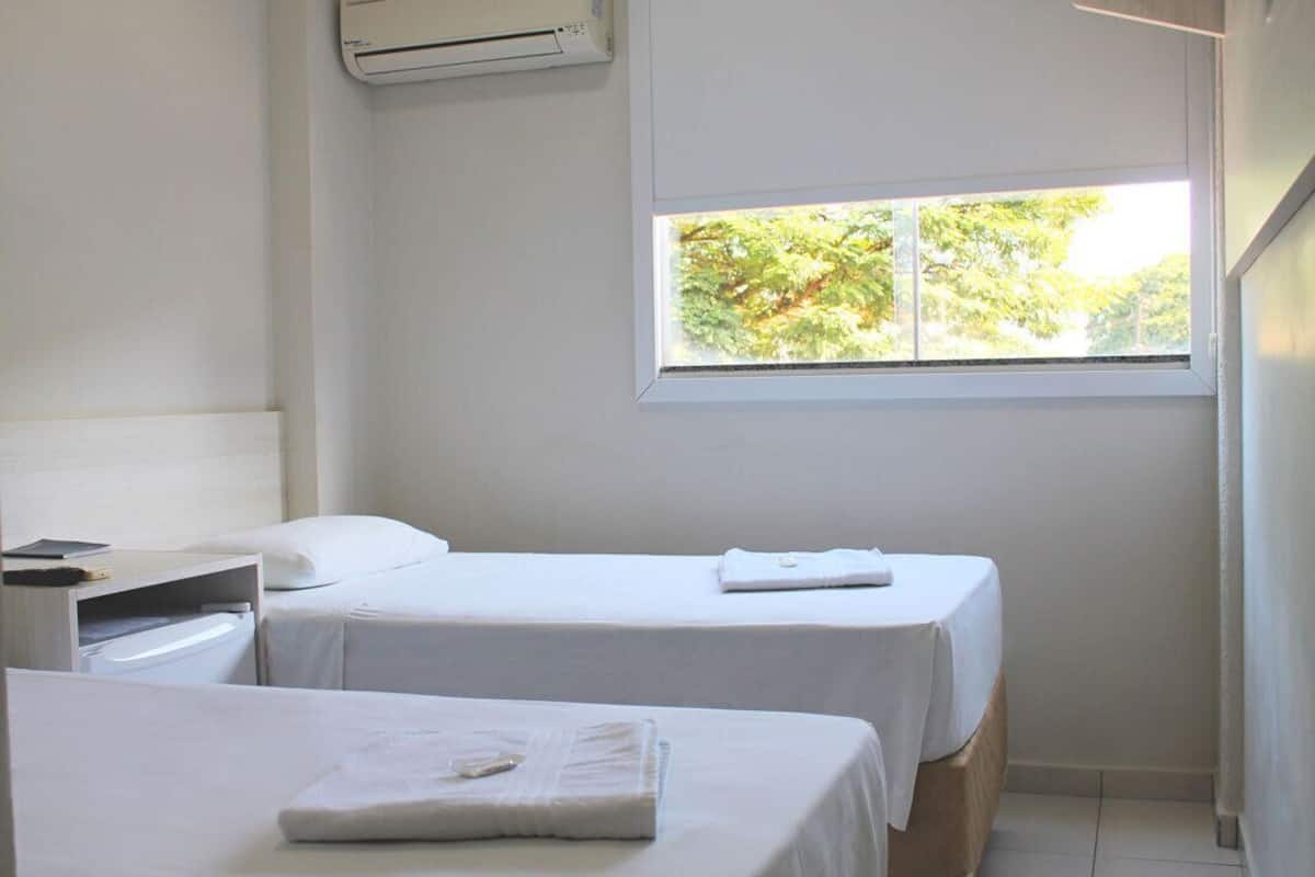 A hotel room with two beds, a window with a view of trees, and an air conditioning unit on the wall.