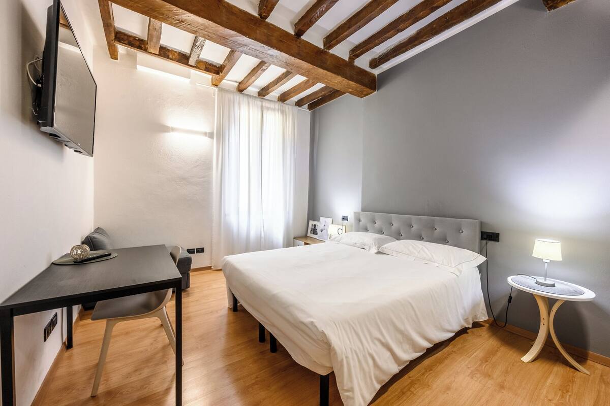 A hotel room with a bed, a desk, a chair, a table lamp, and a TV mounted on the wall.