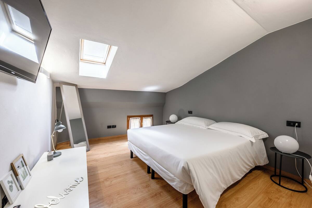A bedroom with a bed, a desk, a mirror, a round table, and a skylight.
